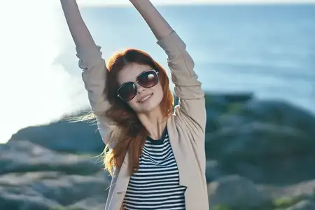 Женщина в отпуске жестикулирует, подняв руки над головой, на фоне моря и гор