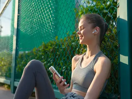Молодая женщина в наушниках отдыхает на улице, держа смартфон, сидя у зелёного забора