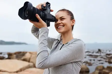 Радостная молодая женщина в сером спортивном топе фотографирует на камеру у моря, воплощая дух outdoor-фотографии и исследования природы