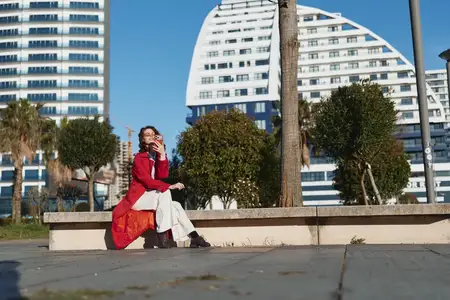 На ней белое платье под пальто, а изогнутые белые здания и пальмы образуют городской фон, создавая расслабленное уверенное настроение