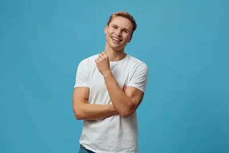 Улыбающийся счастливый молодой мужчина европеоидной расы в белой футболке смотрит в камеру со скрещенными руками, уверенная повседневная поза, изолирован на синем фоне