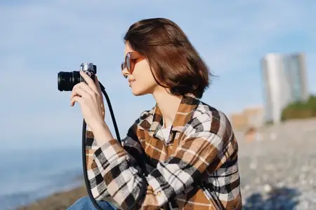 Портрет на открытом воздухе: женщина с фотоаппаратом у моря, в клетчатой рубашке и солнечных очках