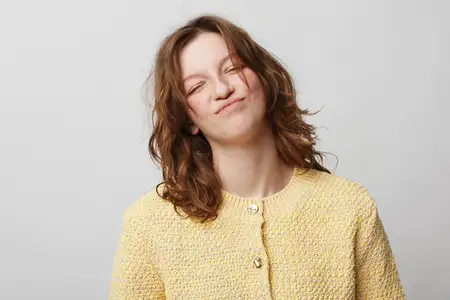 Улыбающаяся кудрявая женщина в желтом свитере демонстрирует игривое выражение лица с закрытыми глазами и поджатыми губами