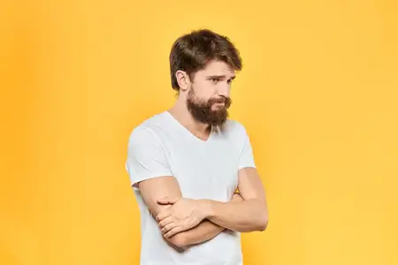 Бородатый мужчина в белой футболке жестикулирует руками, выражая эмоции на студийном жёлтом фоне
