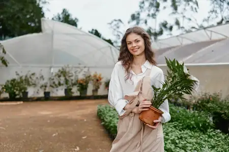 Улыбающаяся молодая женщина в светлой одежде держит горшечное растение в саду на открытом воздухе, демонстрируя связь с природой и устойчивость