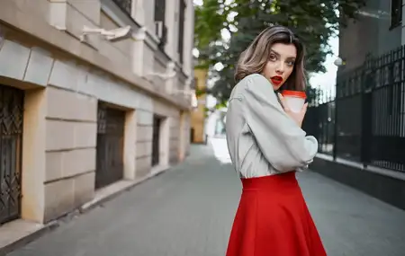 Модная блондинка в красной юбке стоит возле здания, тротуар, парк, природа