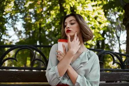 Жизнерадостная женщина сидит на скамейке с чашкой кофе на открытом воздухе в парке