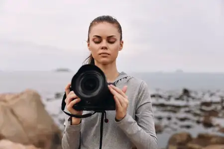 Преданная своему делу молодая женщина-фотограф в серой толстовке, сосредоточенная на своей камере на безмятежной сцене пляжа с каменистым берегом под мягким небом, что идеально подходит для…