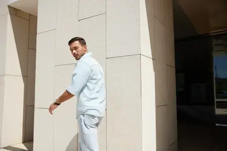 Молодой человек в повседневной светлой одежде проходит мимо современного здания, оборачиваясь с любопытным взглядом