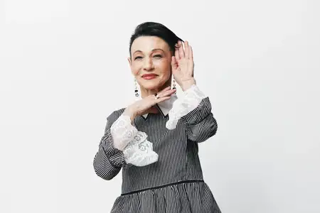 Азиатская женщина в сером платье позирует, закрывая лицо руками перед камерой