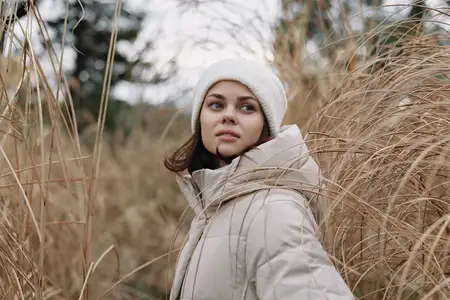 Сцена на открытом воздухе: задумчивая женщина в бежевом пальто и шапке-бини среди высокой травы