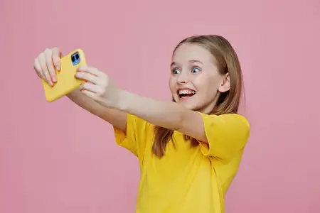 Радостная девушка делает селфи на смартфон на фоне ярко-розового фона, одетая в желтую футболку, излучая счастье и позитив