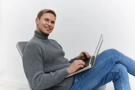 Улыбающийся мужчина работает за ноутбуком, одетый в серую водолазку, сидит в удобном кресле в повседневной одежде на белом фоне, сосредоточенный на образе жизни фрилансера и цифрового кочевника