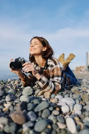 Женщина-фотограф на каменистом берегу с камерой наготове, на открытом воздухе при солнечном свете, в повседневной одежде, с радостным выражением лица снимает пляжные сцены и детали гальки