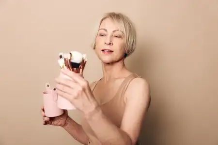 Светловолосая женщина средних лет с короткими волосами выбирает кисти для макияжа из розового держателя в нейтральной студийной обстановке
