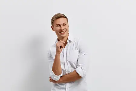 Улыбающийся мужчина, работающий как цифровой кочевник, одетый в белую рубашку, стоит изолированно на однотонном фоне с непринужденным уверенным взглядом, олицетворяя технологии и образ жизни…