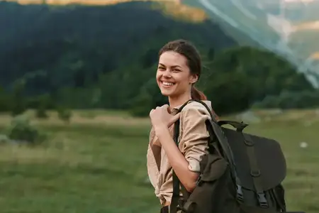 Улыбающаяся женщина с рюкзаком стоит на травянистом поле возле леса и гор, воплощая активный образ жизни и путешествия на свежем воздухе в естественной среде