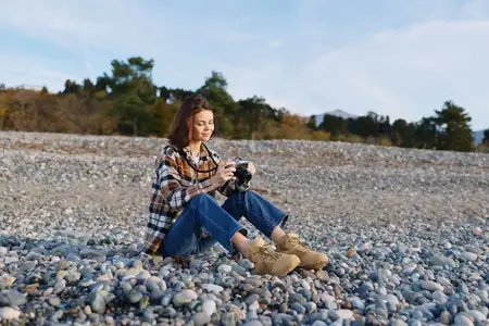 Непринужденная outdoor-съемка с женщиной и камерой на каменистом берегу, на заднем плане безмятежные горы, сосредоточенный фотограф запечатлевает природу и моменты путешествия при дневном свете
