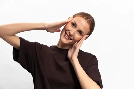 Счастливая женщина с каштановыми волосами в темной футболке широко улыбается и держит руки у лица на чистом белом фоне