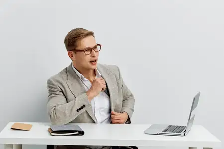 Молодой мужчина европеоидной внешности работает за ноутбуком, разговаривает по видеосвязи, одет в серый клетчатый пиджак и белую рубашку, в очках, сидит за столом на светлом фоне