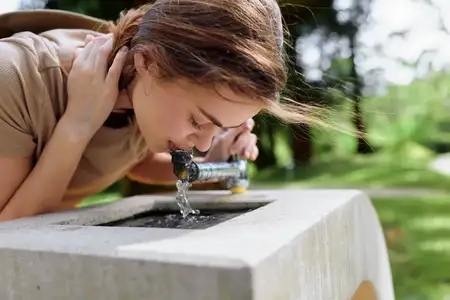 Женщина пьет воду из уличного бетонного фонтана в зеленом парке