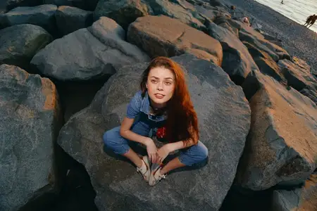 Женщина в стиле лайфстайл, одетая в современный бохо-вестерн деним в уличном стиле, сидит на больших камнях у моря, освещенная теплым естественным светом и кинематографическими цветовыми оттенками