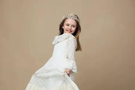 Веселая маленькая девочка в красивом белом кружевном платье и сверкающей короне, ярко улыбаясь, кружится на фоне мягкого светло-коричневого фона