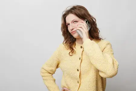 Кудрявая женщина в жёлтом вязаном свитере прижимает смартфон к уху с задумчивым видом