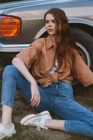 Уверенная молодая женщина в oversized рубашке и джинсах сидит рядом с винтажным автомобилем, демонстрируя сочетание ностальгии и современной моды в земляных тонах, что идеально подходит для…