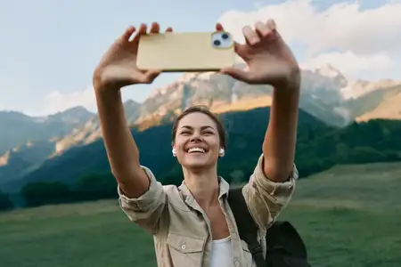 Молодая женщина в легкой куртке и с рюкзаком делает селфи на смартфон на фоне живописных гор под голубым небом с облаками