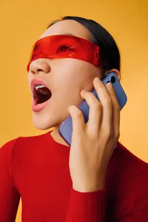 Молодая женщина в красных очках и красном топе держит смартфон у уха, кричит или страстно говорит на желтом фоне