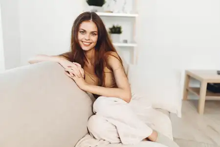 Уютная, счастливая женщина сидит на белом диване и смотрит в камеру