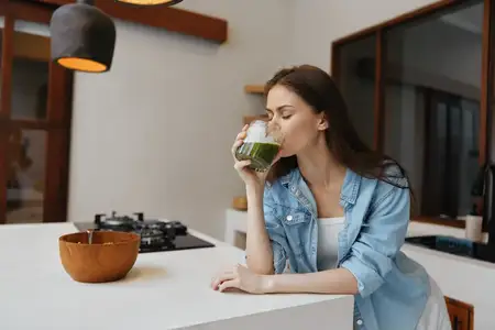Молодая женщина с длинными каштановыми волосами пьет зеленый смузи на современной кухне