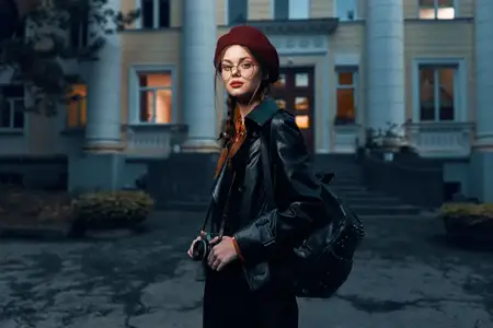 Стильная женщина в красном берете и очках принимает эффектную позу на фоне залитого солнцем исторического здания