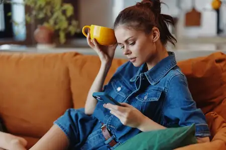 Женщина отдыхает на диване с чашкой кофе, пользуется смартфоном и наслаждается досугом дома