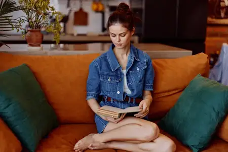 Женщина в джинсовой куртке читает книгу, сидя на оранжевом диване в уютной обстановке гостиной