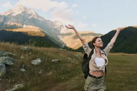 Жизнерадостная женщина с рюкзаком исследует горный ландшафт, стоя в травянистой долине под ясным небом