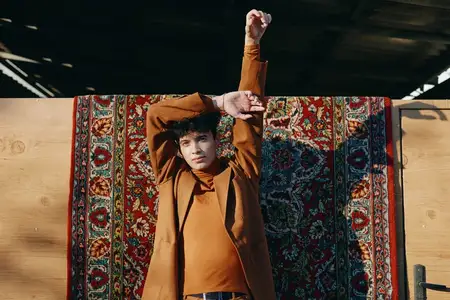 Молодой человек потягивается, стоя перед красивым фактурным ковром, одетый в трендовую повседневную одежду, подчеркивающую современную эстетику моды