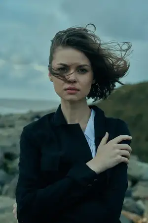 Уличная сцена с фигурой, обдуваемой ветром на побережье, темная блуза, развевающиеся от порывов ветра волосы, штормовые облака, суровый пейзаж, передающие стойкость и напряженную атмосферу