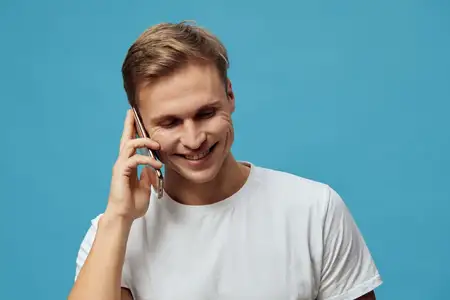 Счастливый молодой человек с короткими каштановыми волосами, одетый в однотонную белую футболку, прижимает мобильный телефон к уху, улыбается и участвует в разговоре на фоне ярко-голубого фона, что…
