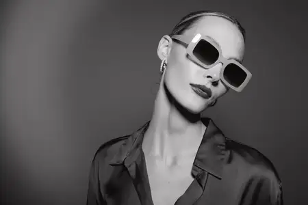 Стильная светлокожая модель европейской внешности в oversized-солнцезащитных очках и шелковой одежде уверенно позирует в черно-белой студийной обстановке с драматичным освещением