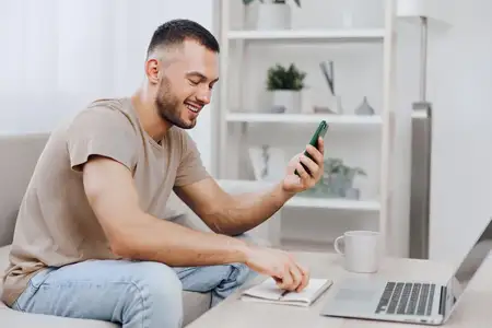 Веселый мужчина пользуется смартфоном, сидя за стильным домашним рабочим местом