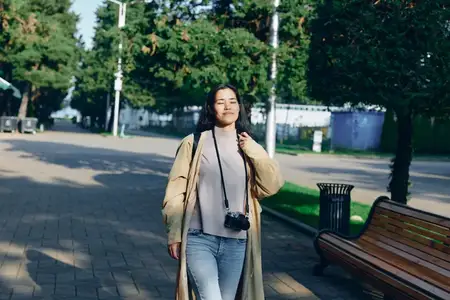 Уличная сцена: женщина в длинном пальто с фотоаппаратом на груди прогуливается мимо деревьев и скамеек в залитом солнцем парке