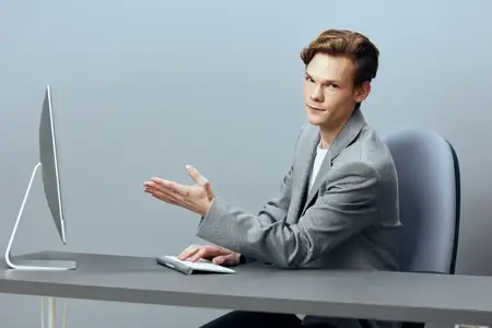 Успешный молодой руководитель сидит за белым столом, уверенно работая за ноутбуком в современном офисе