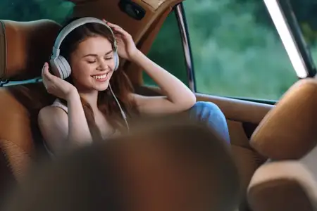 Радостная молодая женщина в наушниках улыбается, слушая музыку в автомобиле, в окружении яркой зеленой природы, что подчеркивает расслабление и счастье в повседневном летнем наряде