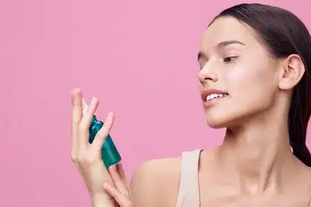 Красивая молодая женщина с сияющей улыбкой использует зеленую сыворотку на фоне мягкого розового фона, подчеркивая естественную красоту, процедуры по уходу за кожей и темы благополучия