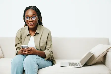 Стильная афроамериканская женщина, бизнес-фрилансер, работающая сидя на диване дома с ноутбуком и телефоном, деловые звонки и переписка, домашняя одежда и очки, светлый интерьер на заднем плане