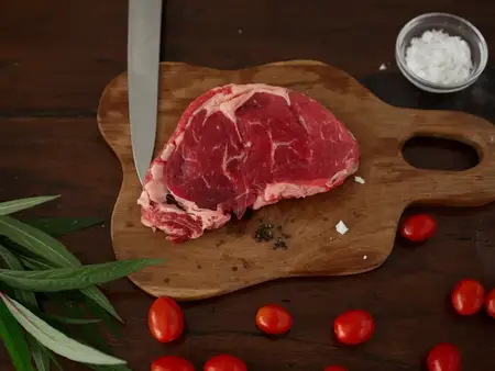Композиция из сырого стейка на деревянном кухонном столе со специями, солью, овощами и зеленью, процесс приготовления, вид сверху, свободное место для рецептов