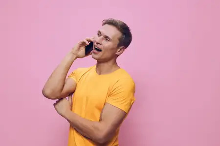 Счастливый мужчина в жёлтой футболке пользуется мобильным телефоном, улыбается и наслаждается разговором, стоит на розовом фоне, повседневная одежда, весёлая и расслабленная атмосфера, дизайн образа…