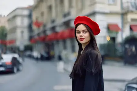 Портрет модной красивой женщины, стоящей на улице на фоне города в стильной одежде, с красными губами и красным беретом